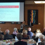 A woman speaks behind a podium and in front of a screen that says "Opera Grand Rapids Future", while a dozen people are seated and listening